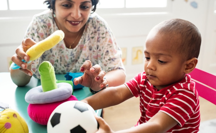 Toddler boy plays with female teacher and toys at a table.