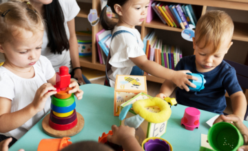 Three toddlers play with stacking toys and blocks in classroom as teacher looks on.