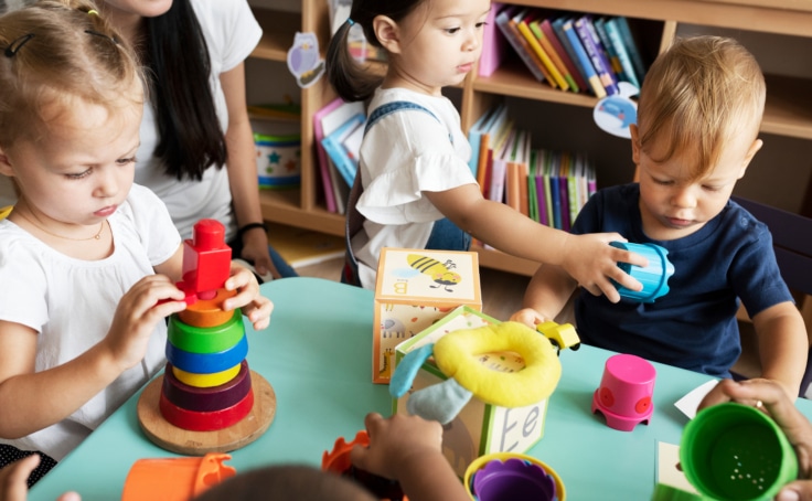 Three toddlers play with stacking toys and blocks in classroom as teacher looks on.