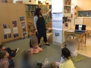 Teacher instructing preschoolers who are seated on a rug.
