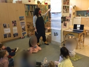 Teacher instructing preschoolers who are seated on a rug.