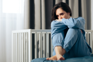 Sad woman sits with arms crossed on knees on the bed, with empty crib in background.