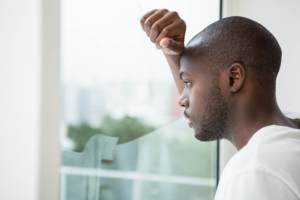 Black man gazes through window with hand on head.