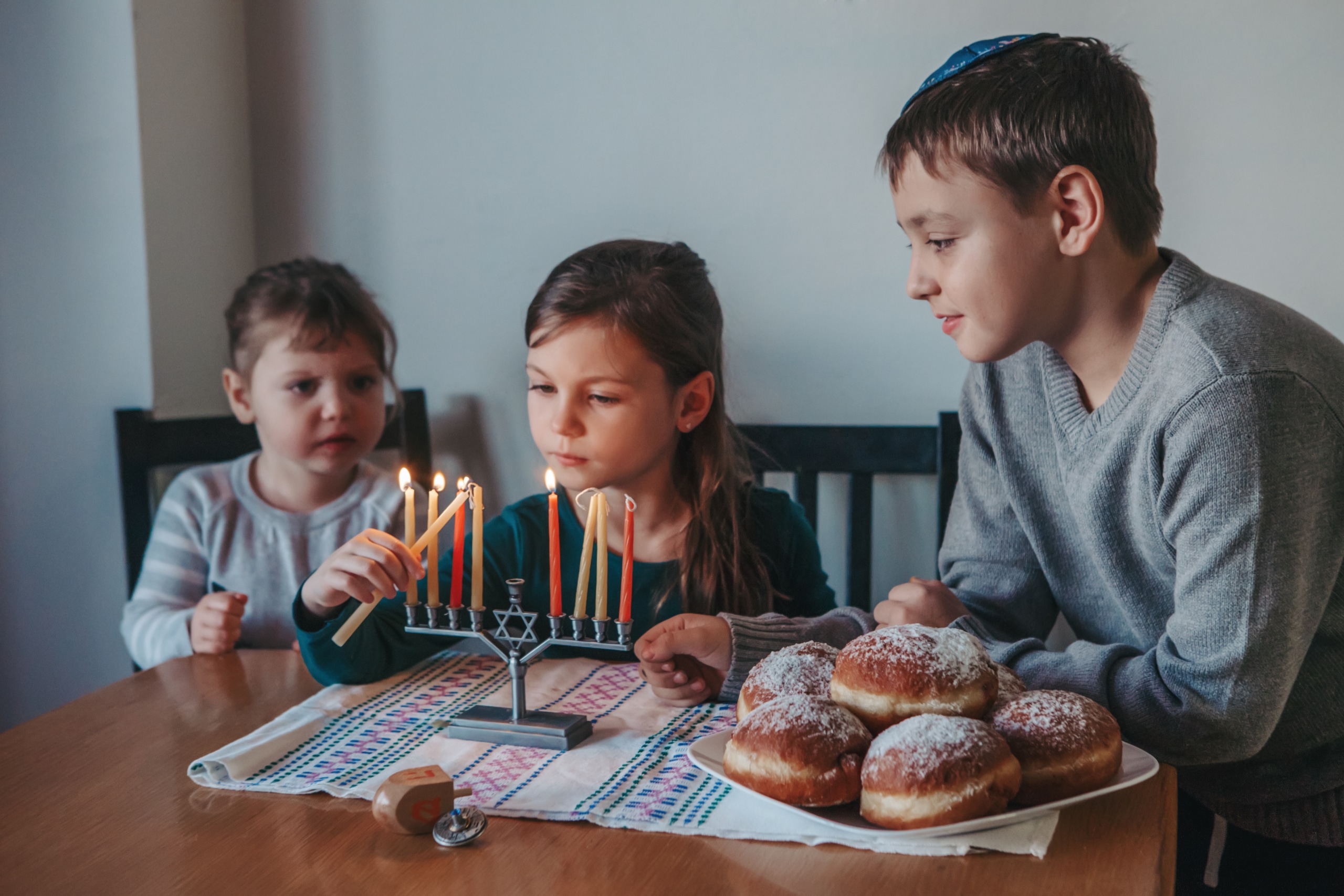 Brother and sisters siblings lighting candles on menorah for Jewish Hanukkah holiday at home. Children celebrating Chanukah festival of lights. Dreidel and Sufganiyot donuts in plate on a table.