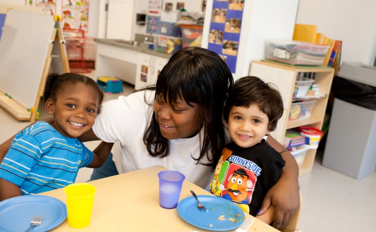 A child care worker sits at a table with two children as they enjoy breakfast