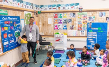 A teacher leads a room full of toddlers in front of a blue board