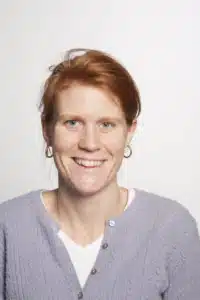 a smiling woman with short red hair wearing a white shirt and light purple sweater