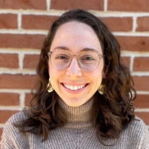 a woman with long brown hair wearing eyeglasses and smiling, wearing a light brown turtleneck sweater