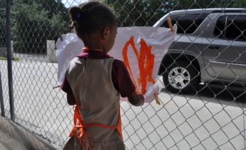 a child wears an apron and stands behind a fence with a drawing