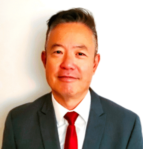 a smiling man with short dark hair wearing a dark grey suit jacket with white shirt and red tie