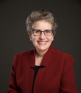 a smiling woman with short light brown hair and glasses wearing a red blazer with black shirt
