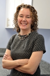 a smiling woman with curly sandy blonde and brown hair wearing a grey and black striped blouse with her arms crossed