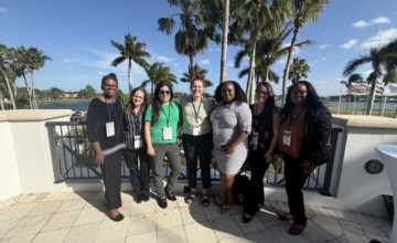 a group of woman standing close together on a landing with a body of water and palm trees in the background