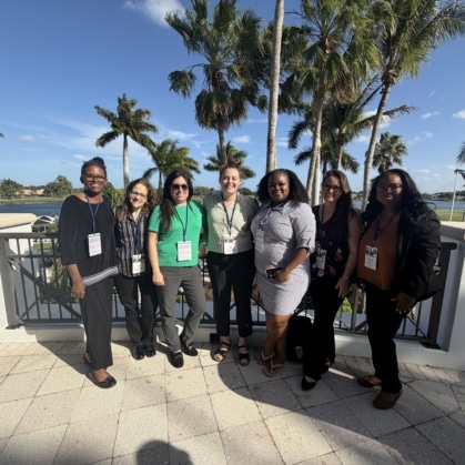 a group of woman standing close together on a landing with a body of water and palm trees in the background
