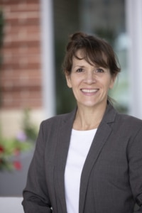 a smiling woman with brown pulled up on top of her head wearing a dark grey blazer with white shirt