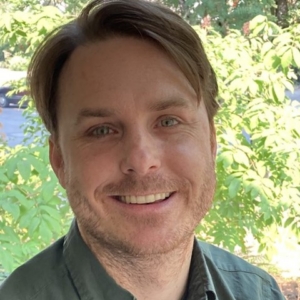 a man with short light brown hair smiling and wearing a dark green shirt