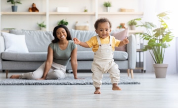 a young male child learns to walk in the foreground with mother sitting on the floor behind him