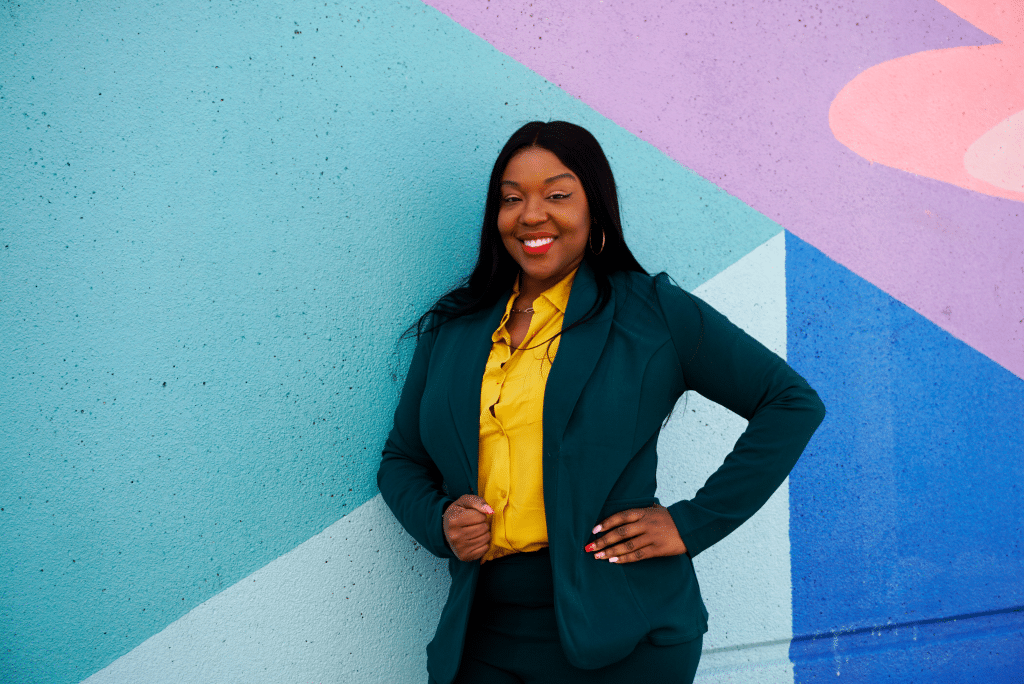 a smiling woman with long black hair wearing a yellow dress shirt and navy blue blazer stands against a multi-colored wall