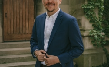 a smiling man with short dark hair wearing a white shirt with navy suit