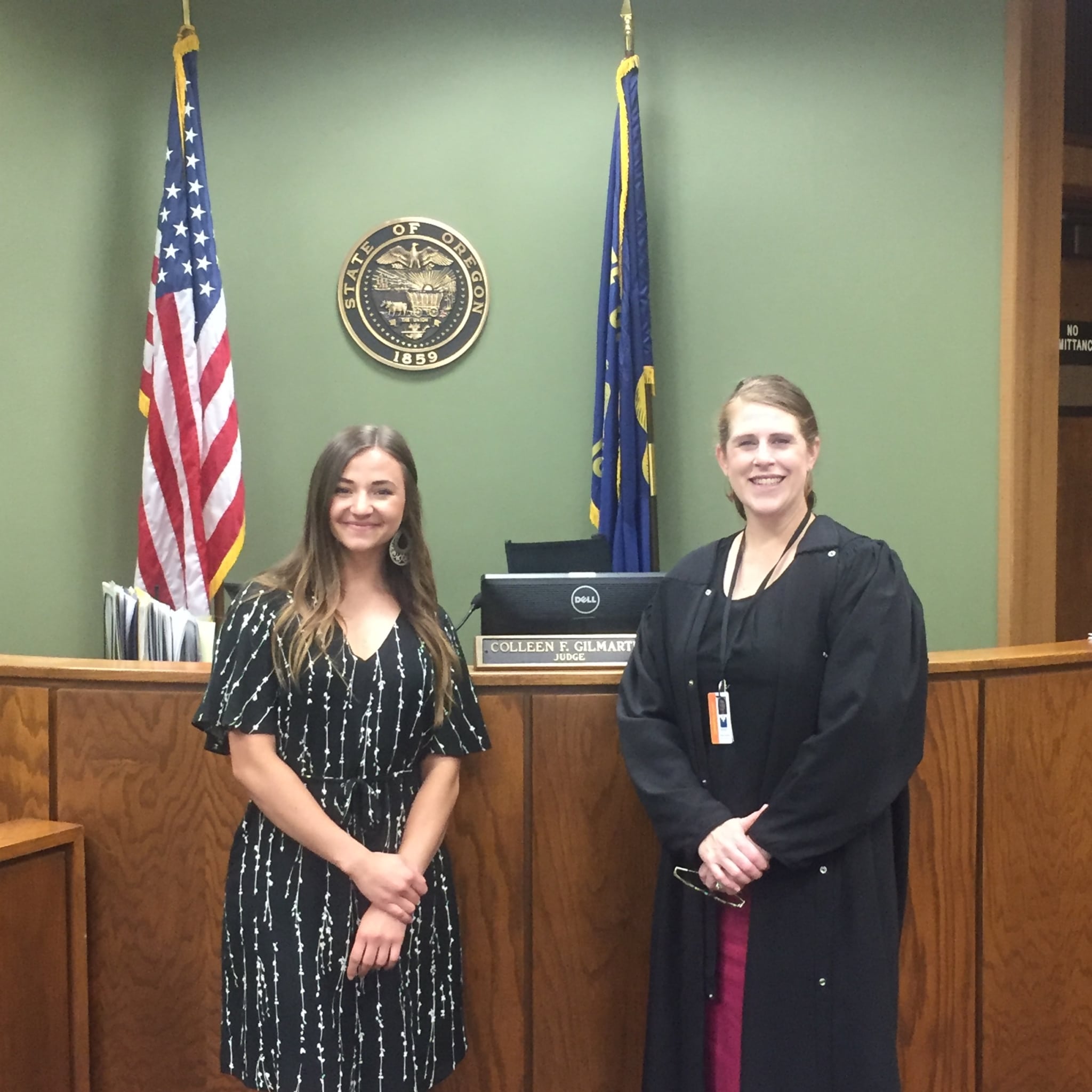 two smiling women standing in courtroom in front of a judge's desk, one of the women is a judge