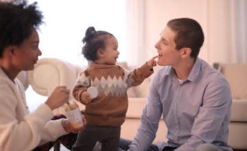 a toddler points at father's mouth while mother holds a snack looking on