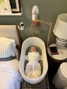 A smiling baby wrapped in a sleep sack inside of a Snoo-brand bassinet. 