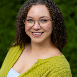 a smiling woman with shoulder-length curly hair wearing glasses, a white undershirt, and neon green button up sweater