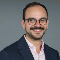 a smiling man with short dark hair wearing glasses and a pink button up shirt with navy suit jacket