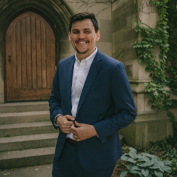 a smiling man with short dark hair wearing a white shirt with navy suit