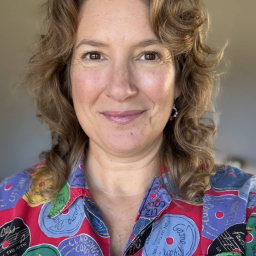 a smiling woman with light brown hair wearing a multi-colored button up shirt